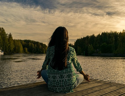 A woman meditating at a lake peacefully. She has brown hair, a green dress, and is sitting by the water.